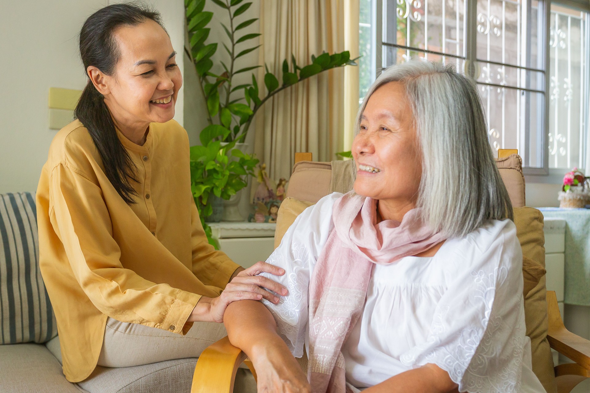 Asian mature woman talking and holding hand with senior woman sitting on wheelchair in living room at nursing home